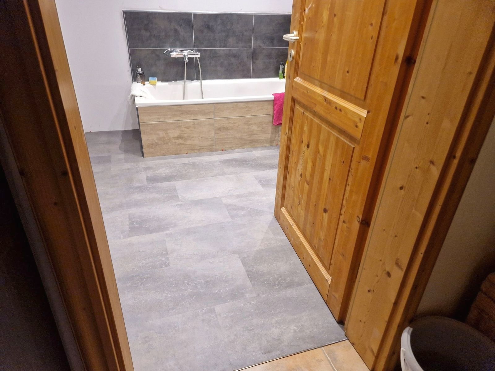 Bad mit grauem Vinylboden in Steinoptik, im Hintergrund eine Badewanne, vorne eine Holztür