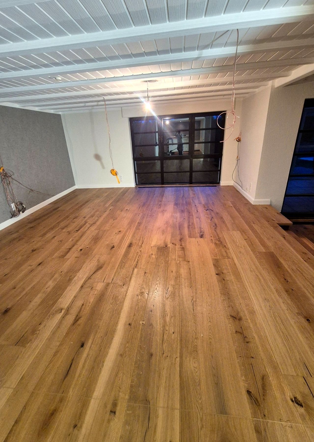 Großer Raum mit langen Eichendielen bis zu einer schwarzen Sprossen-Glastür, darüber eine weiß gestrichene Holzdecke