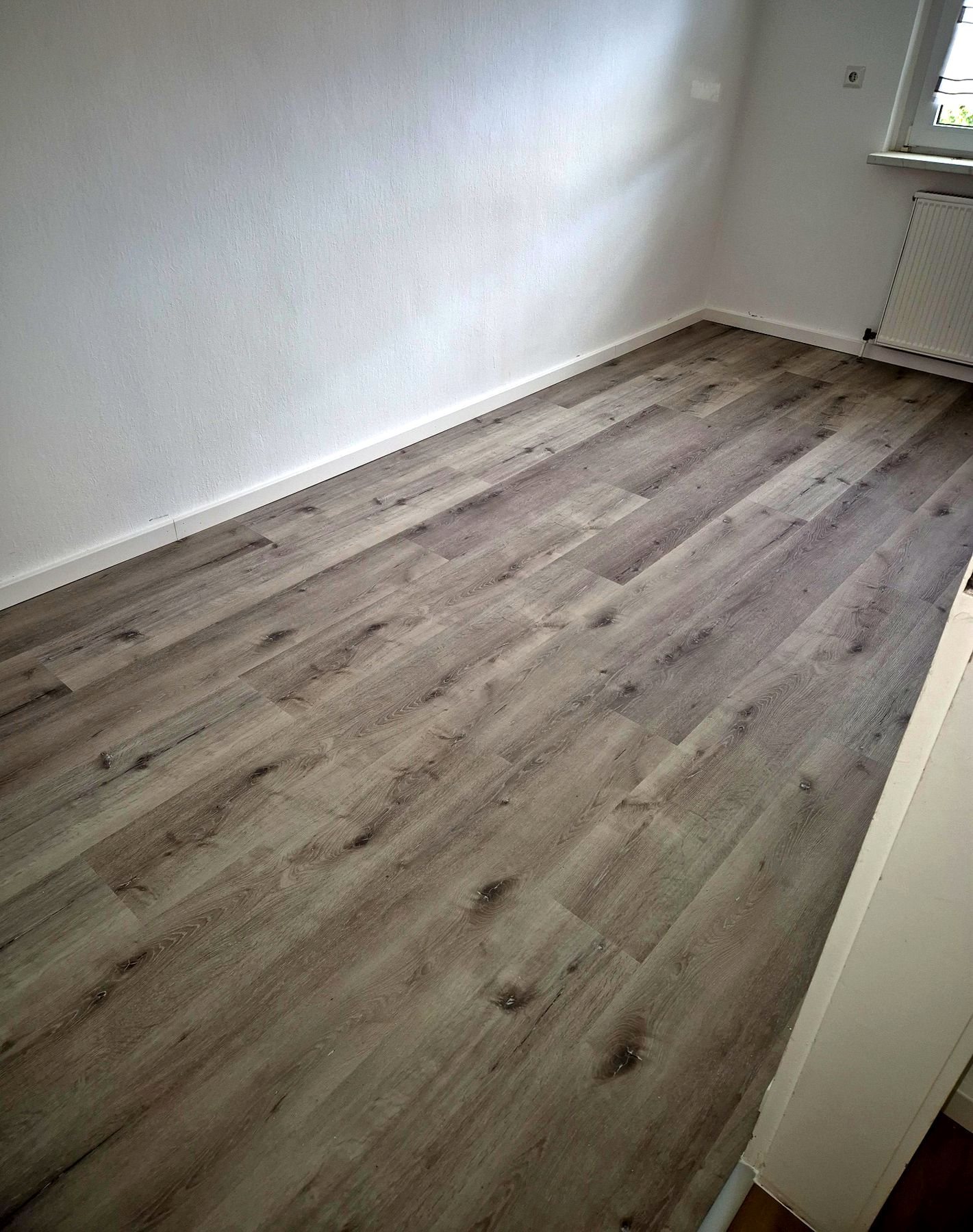 Leeres Zimmer mit grauem Vinylboden in Dielenoptik und weißen Sockelleisten